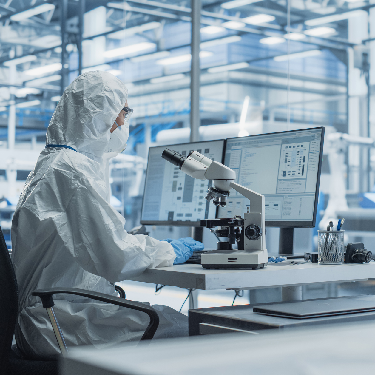 modern factory: industrial engineer working on desktop computer in clean sterile coveralls. scientist using a microscope, developing advanced solutions for high tech medical vaccine and gene research